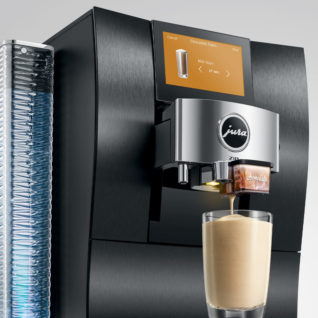 A sleek black and stainless steel Jura coffee machine with a digital display screen, shown dispensing a latte into a glass. A textured water tank is visible on the left, all set against a neutral light gray background.