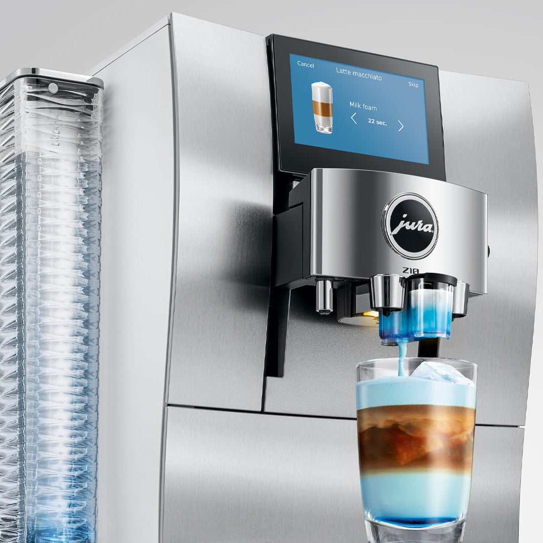 A stainless steel Jura coffee machine with a digital touch screen and a glass of layered coffee topped with blue foam, shown against a light gray background.